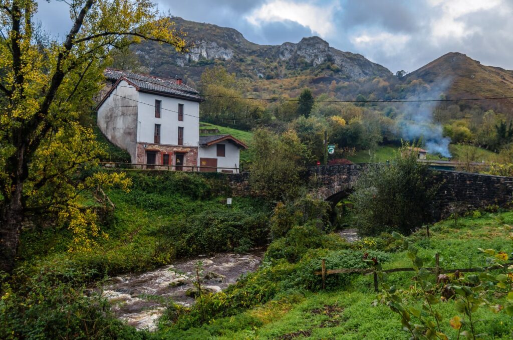 Casa junto al río con puente de piedra y montañas